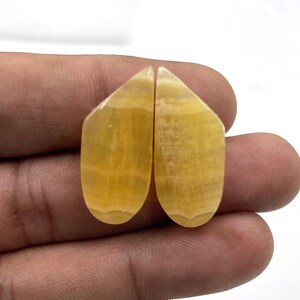 Può includere: Due pietre preziose di calcite gialla lucidata, con un aspetto a strati e venato. Le pietre sono tenute in una mano, mostrando le loro forme lisce e curve e le variazioni di colore naturali. Ideale per la creazione di gioielli.