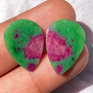 May include: Two teardrop-shaped gemstones. Each stone has a vibrant green base color with a prominent, irregular pattern of deep magenta hues. The stones are smooth and polished, reflecting light.
