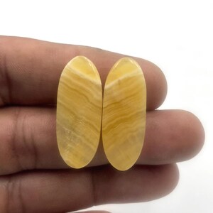 Può includere: Due pietre preziose ovali e lucide, di colore giallo caldo e con un motivo a bande. Le pietre sono tenute in una mano, mostrando la loro superficie liscia e lucida. Le pietre sembrano essere un tipo di onice o agata.