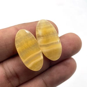 Può includere: Due pietre preziose ovali lucide, con un motivo a strati e a bande nei toni del giallo e del crema. Le pietre sono tenute in una mano, mostrando le loro superfici lisce e lucide. Queste pietre sono ideali per la creazione di gioielli.