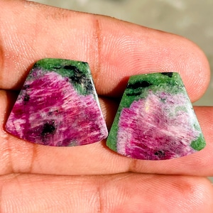 Könnte beinhalten: Zwei polierte Edelstein-Cabochons. Jeder Stein zeigt ein lebendiges rosa-weißes Muster mit grünen und schwarzen Akzenten. Die Steine sind trapezförmig, ideal für die Schmuckherstellung oder das Sammeln.