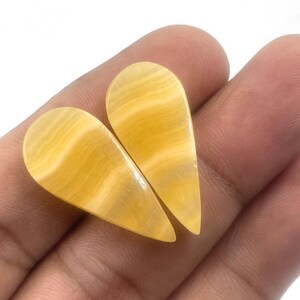 Può includere: Due gemme a forma di goccia con un motivo a strisce gialle e bianche. Le pietre lucide sono tenute in una mano, mostrando le loro superfici lisce e riflettenti. Le gemme sono di colore giallo chiaro.