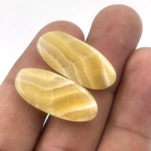 Può includere: Due pietre preziose ovali lucide, di colore giallo caldo con fasce bianche. Le pietre sono tenute in una mano, mettendo in risalto le loro superfici lisce e lucide. Le pietre hanno un aspetto naturale e organico.