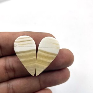 Può includere: Due pietre preziose a forma di cuore con un motivo a bande crema e beige. Le pietre sono tenute in una mano, mostrando le loro superfici lucide e le variazioni di colore naturali. Cabochon per la creazione di gioielli.