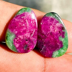 Pode incluir: Duas pedras preciosas polidas em forma de gota. As pedras apresentam um vibrante centro magenta, circundado por uma faixa verde. As gemas têm uma aparência natural e texturizada, com veios visíveis e variações de cor.