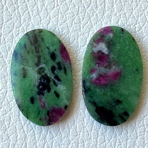 Può includere: Due pietre preziose di zoisite di forma ovale. Le pietre hanno un colore di base verde con inclusioni nere scure e magenta vibranti. Le pietre preziose sono lucide e lisce, con un aspetto naturale e organico. Le pietre sono poste su uno sfondo neutro.