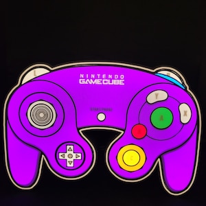 May include: A purple Nintendo GameCube controller with white buttons and a black background. The controller has a circular button in the center, a directional pad, and four face buttons labeled A, B, X, and Y.