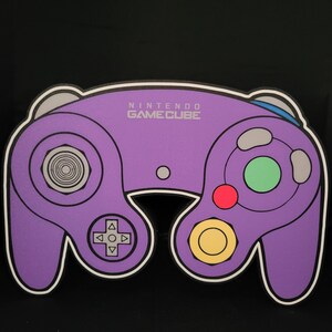 May include: Purple Nintendo GameCube controller with a white outline. The controller has a circular button in the center, a directional pad, and two analog sticks. The text "NINTENDO GAMECUBE" is printed in white on the controller.
