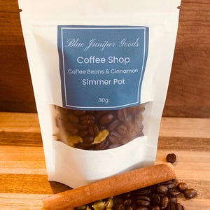 May include: A white pouch with a clear window showing coffee beans, cardamom pods, and a cinnamon stick. The pouch has a blue label with the text "Blue Juniper Goods Coffee Shop Coffee Beans & Cinnamon Simmer Pot 30g". The items are on a wooden surface.
