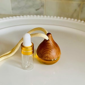 May include: A wooden car air freshener with a clear glass bottle filled with essential oil. The bottle has a gold cap and a dropper. The air freshener is attached to a white string.