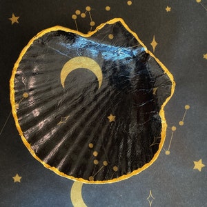 May include: A black seashell with a gold rim and a gold crescent moon design on a black background with gold stars and constellations.