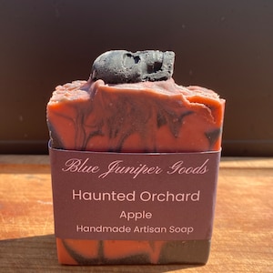 May include: Artisan soap bar with a dark red and black swirl design, topped with a small black skull. The soap is wrapped with a brown label that reads "Blue Juniper Goods Haunted Orchard Apple".