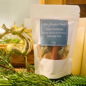 May include: A white pouch labelled "Blue Juniper Goods" with "Yule Solstice" text, containing a simmer pot with orange, spruce, and cinnamon. A gold reindeer figurine and green pine branches are in the foreground. The simmer pot weighs 30g.