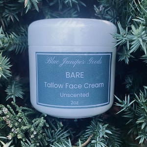 May include: A white jar of "Bare Tallow Face Cream" from "Blue Juniper Goods" is surrounded by green foliage. The teal label has white text, stating the product is unscented and contains 57g.