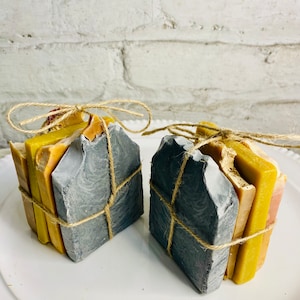 May include: Two sets of handmade soap bars wrapped in twine and tied with a bow. The soap bars are layered in shades of yellow, orange, and brown. The soap bars are wrapped in a dark grey material.