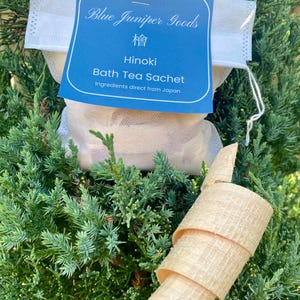 May include: A Blue Juniper Goods bath tea sachet with a blue label, reading "Hinoki Bath Tea Sachet, Ingredients direct from Japan." The white sachet is set against green foliage, with a rolled piece of light-coloured wood in the foreground.