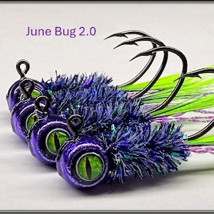 May include: Several fishing lures with the text "June Bug 2.0". The lures have purple, blue, and green accents, with large, curved black hooks. The eyes of the lures are green.