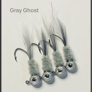 May include: Four gray and silver fishing jig heads with iridescent eyes and white and gray feathers. The jig heads are labeled "Gray Ghost".