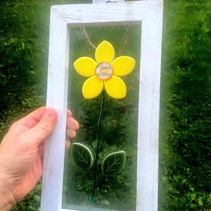 May include: A framed floral decoration featuring a yellow flower with a center that reads "Watch us BLOOM." The flower has green leaves and a dark stem. The frame is white with a distressed finish, and the background is a blurred green.
