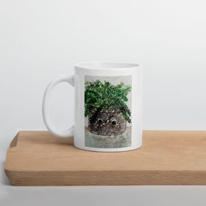 May include: A white ceramic mug featuring a printed watercolor illustration of a dark brown, textured plant pot with two large, round, white-and-black googly eyes. The pot contains a lush, vibrant green fern with detailed, leafy fronds.