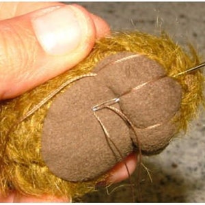 Op de afbeelding: Close-up van een bruine teddybeerpoot die dichtgenaaid wordt met een naald en draad. De poot is gemaakt van bruin stof en bedekt met gele vacht.