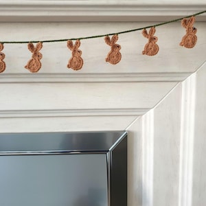 May include: A decorative garland featuring crocheted brown bunny rabbits suspended from a green cord. The bunnies are evenly spaced, ideal for Easter or spring celebrations. The garland is a charming handmade decoration.