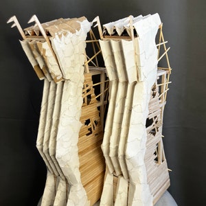 May include: A model of a building made from cardboard and wooden sticks. The building is white and has a textured surface. The building is in a state of disrepair, with some of the walls missing and the roof caved in.