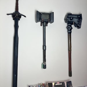 Puede incluir: Tres armas de estilo medieval colgadas en una pared blanca. A la izquierda, una espada larga y negra con empuñadura marrón. A la derecha, dos martillos grises oscuros con mangos marrones, uno con cabeza cuadrada y otro con cabeza redondeada.