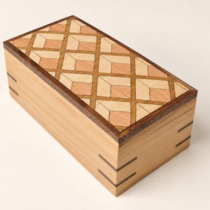 Può includere: Una scatola rettangolare in legno con un coperchio caratterizzato da un motivo geometrico a intarsio in tonalità di legno chiare e medie. Il corpo della scatola è in legno chiaro con giunzioni angolari scure e una cornice scura.