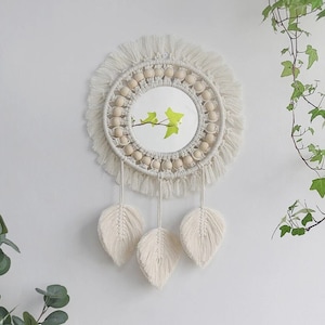 May include: A white macrame wall hanging with a round mirror in the center. The mirror is surrounded by wooden beads and the hanging has three white feather-shaped tassels.