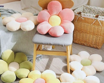 May include: Five plush daisy-shaped floor cushions in different colors: pink, white, green, yellow, and cream. The cushions are arranged on a wooden floor and a wooden chair.
