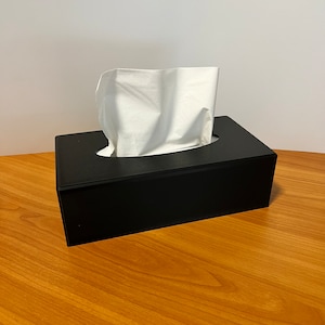 Op de afbeelding: Een zwarte tissueboxhouder met een witte tissuebox erin. De tissueboxhouder staat op een houten oppervlak.