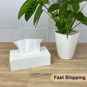 Puede incluir: Un dispensador de pañuelos de papel blanco con una caja de pañuelos de papel blanca y un pañuelo de papel blanco dentro. La caja de pañuelos de papel está sobre una superficie de madera. Una planta verde en una maceta blanca está en el fondo. El texto "Envío rápido" es visible en la esquina inferior derecha de la imagen.