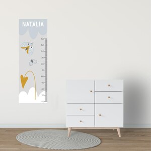 Può includere: Un'asta metrica grigia con illustrazioni di farfalle e cuori, con il nome "NATÁLIA" in alto. L'asta metrica misura fino a 160 cm. Un comò bianco con pomelli e gambe in legno è accanto.