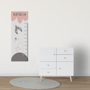 Può includere: Una cassettiera bianca con pomelli e gambe in legno è accanto a un'asta metrica. L'asta metrica è grigia con una parte superiore rosa e presenta il nome "NATÁLIA" e disegni decorativi floreali e a farfalla. L'asta metrica misura fino a 160 cm.