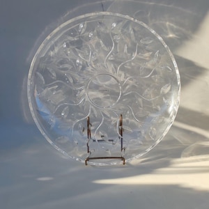 May include: A clear glass serving platter with a leaf design. The platter is on a gold metal stand.