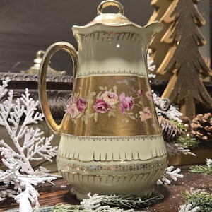 May include: An antique cream-colored porcelain pitcher with a gold handle and trim. The pitcher features a gold band with pink and purple floral designs. A gold lid and a decorative base complete the design. The pitcher is a decorative item.