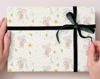 Könnte beinhalten: Eine rechteckige Geschenkbox, verpackt in gemustertem Papier mit Cartoon-Mäusen, Sonnen und floralen Elementen. Die Box ist mit einem schwarzen Band und einer dunkelgrünen Samtschleife befestigt. Das Farbschema ist weich und pastellfarben.