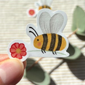 Könnte beinhalten: Ein gestanzter Aufkleber mit einer Cartoon-Biene mit gelben und schwarzen Streifen, durchscheinenden grauen Flügeln und zwei Fühlern. Die Biene befindet sich neben einer kleinen roten Blume mit gelber Mitte. Ein zweiter Aufkleber ist im Hintergrund.