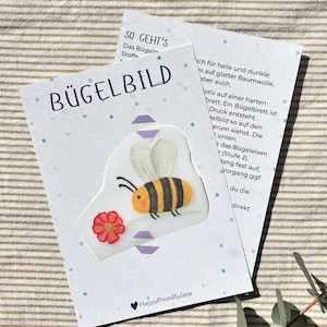 Könnte beinhalten: Eine weiße Karte mit dem Wort "BÜGELBILD" und einem aufbügelbaren Aufnäher in Bienenform. Die Biene ist gelb und schwarz mit weißen Flügeln, und eine rote Blume befindet sich daneben. Die Karte enthält deutschen Text.