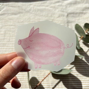 Könnte beinhalten: Ein durchscheinender Aufkleber mit einer Aquarellillustration eines hellrosa Schweins mit einfachem lächelndem Ausdruck, spitzen Ohren und einem geringelten Schwanz, auf einer transparenten Trägerfolie mit Konturschnitt.