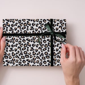 Könnte beinhalten: Rechteckige Geschenkbox mit Leopardenmuster in Schwarz und Beige auf weißem Papier. Sie ist mit einem schwarzen Band und einer dunkelgrünen Samtschleife gebunden. Die Box wird von zwei Händen gehalten.