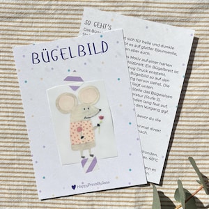Könnte beinhalten: Eine Karte mit dem Wort "BÜGELBILD" in Blau, mit einer Cartoon-Maus, die eine Blume hält. Die Maus trägt ein rosa Hemd mit Herzmustern. Die Karte liegt auf einer gestreiften Oberfläche mit einer zweiten Karte mit Text.