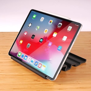 May include: A silver tablet with a white stylus, displayed on a black stand. The screen displays a colorful interface with app icons. The tablet is positioned on a light brown wooden surface.
