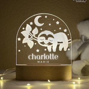 May include: A personalized night light featuring a sleeping sloth design, a crescent moon, and stars. The name "charlotte marie" is illuminated in white. The light sits on a wooden base and is surrounded by fairy lights.