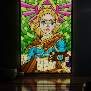 Könnte beinhalten: Buntglas-Kunstwerk mit einer Figur aus The Legend of Zelda. Das Bild zeigt eine blondhaarige Figur in einem blau-braunen Outfit, die ein Gerät hält. Der Hintergrund ist ein Mosaik aus grünem und rosa Glas, schwarz umrahmt.