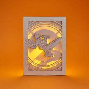 May include: A white paper art piece featuring a detailed outline of a character from the video game Overwatch, illuminated from behind with a warm yellow light. The character is in a dynamic pose, set against a circular Overwatch logo.