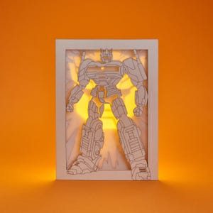 May include: A white paper art piece featuring a robot design, illuminated from behind with a warm yellow light. The robot is depicted in a standing pose within a rectangular frame, set against an orange background. The artwork showcases intricate cut-out details.