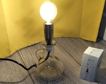 May include: A clear glass lamp base with a round bulb and a black cord. The lamp is on a wood floor and there is a box of LED filament bulbs in the background.
