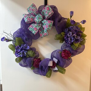 Può includere: Una ghirlanda decorativa con una base in rete viola, adornata con fiori viola e bordeaux, foglie verdi e un fiocco a fantasia floreale. Adatta per la decorazione di porte o pareti.